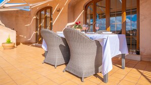 Een terras met een eettafel voor twee personen, met rieten stoelen, een witte tafelkleed en wijnglazen.