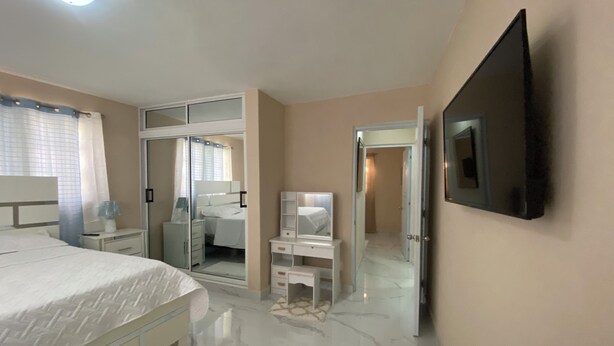 Un dormitorio con cama, vestidor, tocador con espejo y televisor montado en la pared.