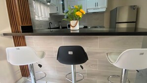 Una cocina moderna con armarios blancos, un refrigerador de acero inoxidable y una encimera negra. Hay dos taburetes blancos para bar y un jarrón con flores en la encimera.