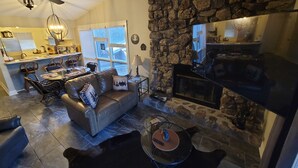 Una sala de estar con chimenea de piedra, un sofá de cuero, un comedor con mesa y sillas, y una cocina al fondo.