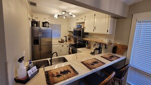Una cocina con armarios blancos, un refrigerador de acero inoxidable, horno empotrado, cafetera y una barra con taburetes.
