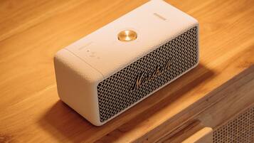 A portable speaker on a wooden surface with a woven lamp overhead.