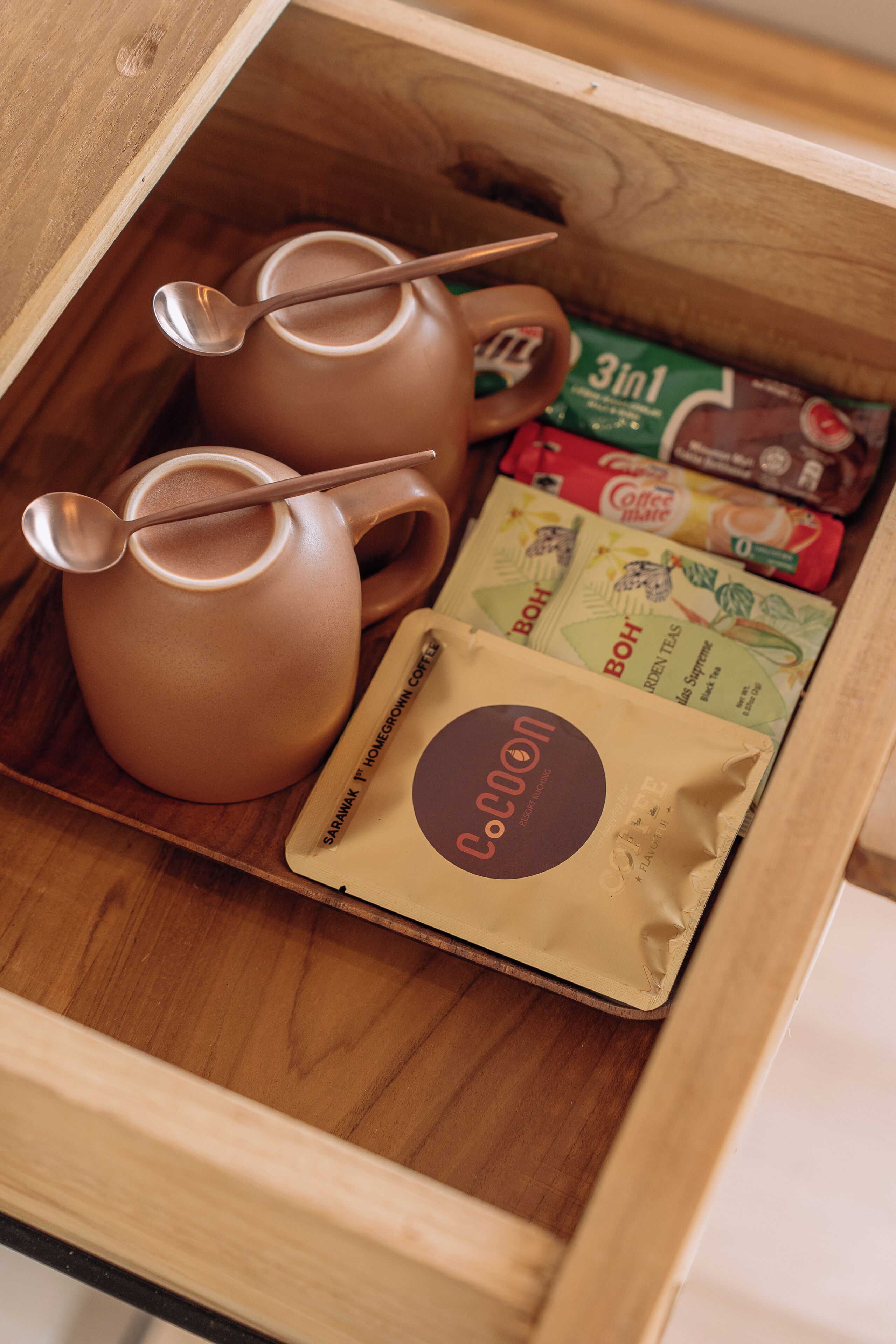 A wooden drawer containing a brown ceramic teapot, two spoons, and several coffee packets.