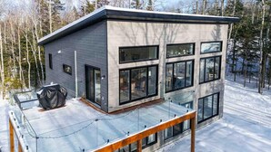 En modern stuga i två våningar med stora fönster, en inglasad veranda och ett snötäckt landskap.