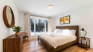 Ett sovrum med en säng i trä, ett skrivbord, en stol, en lampa och ett fönster med utsikt över träd.