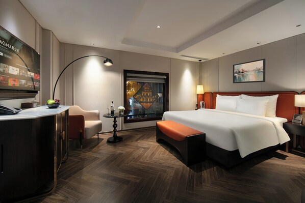 A modern hotel room with a large bed, a desk, a chair, a small table with a lamp, and a painting on the wall.
