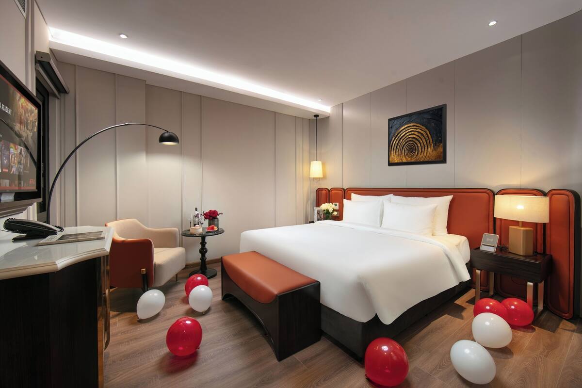 A hotel room with a large bed, a desk with a telephone, a chair, a small table with flowers, and balloons on the floor.