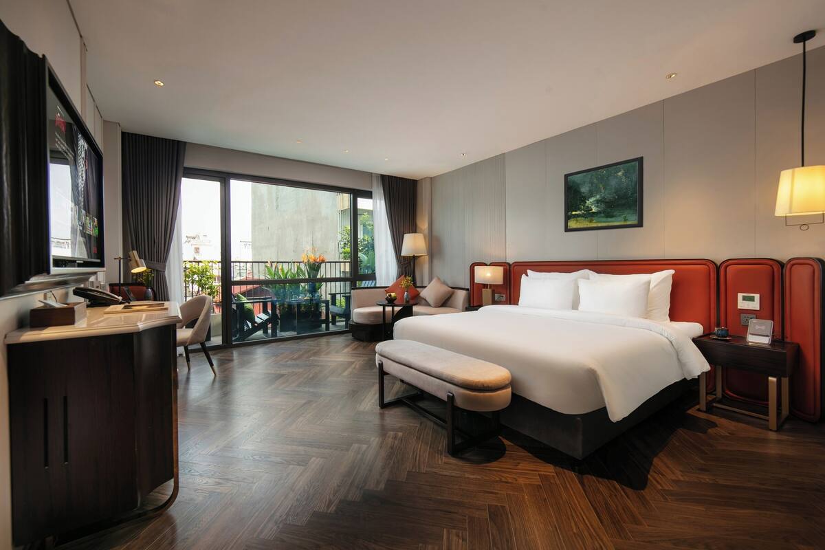 A hotel room with a large bed, a desk, a chair, a bench, a nightstand, a lamp, and a view of the outdoors.