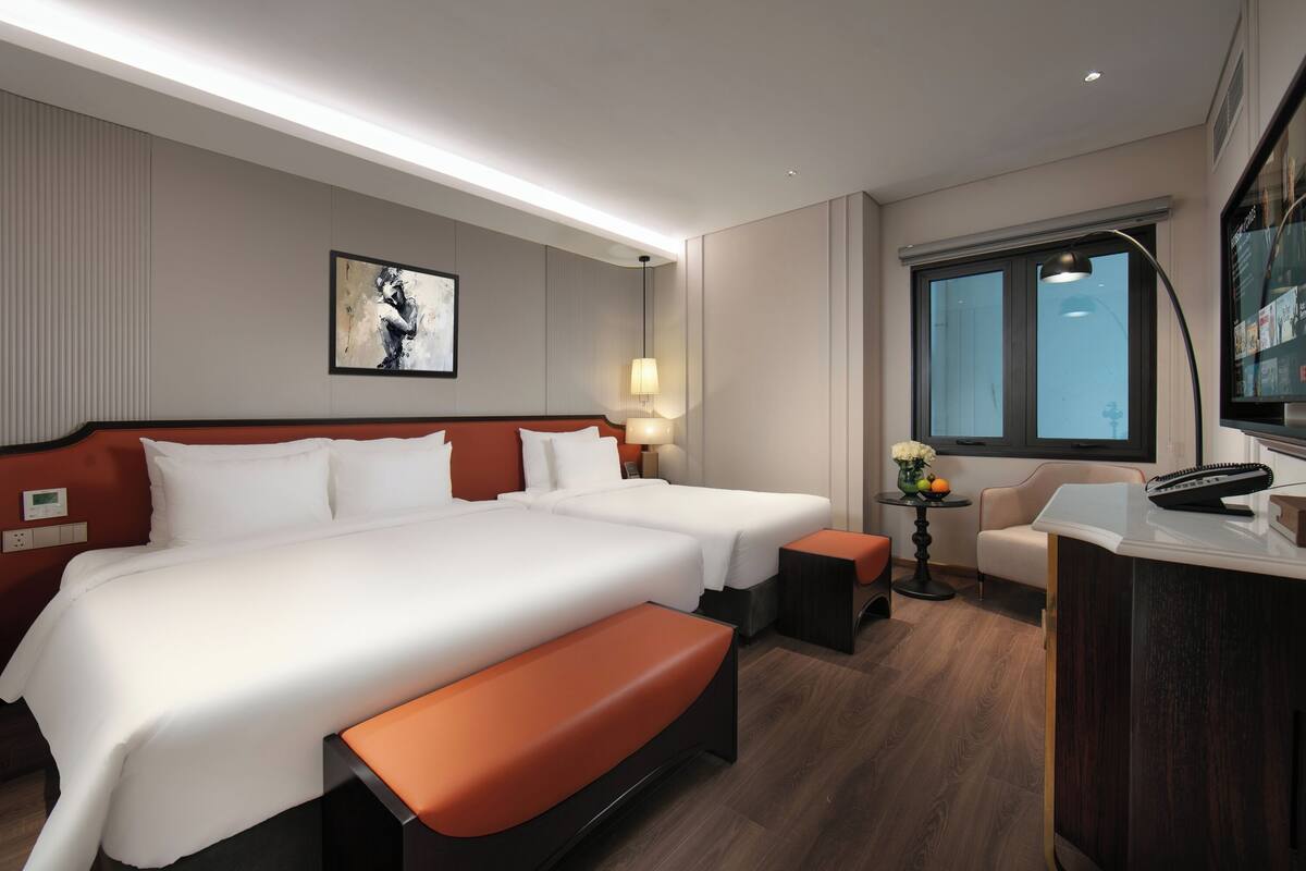 A hotel room with two beds, a desk, a chair, a lamp, a painting on the wall, and a window with a view.