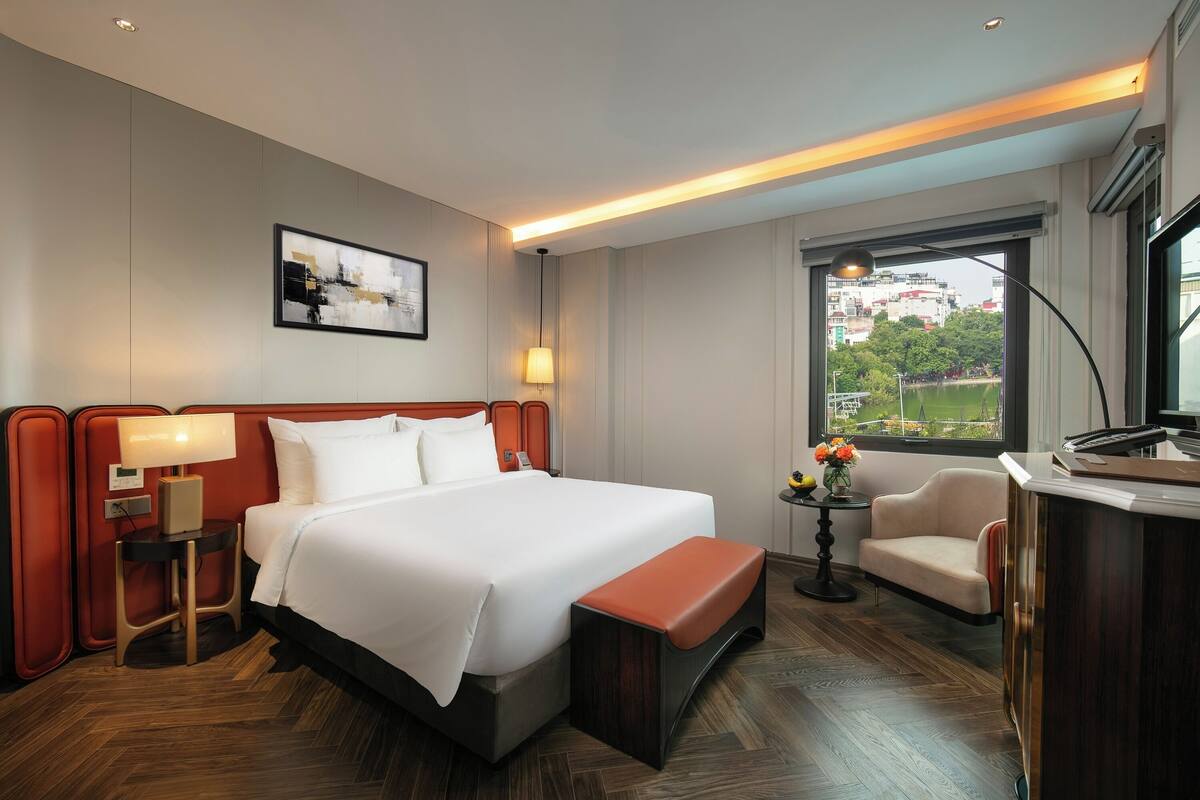 A hotel room with a large bed, a desk, a chair, a small table with flowers, and a window with a view.