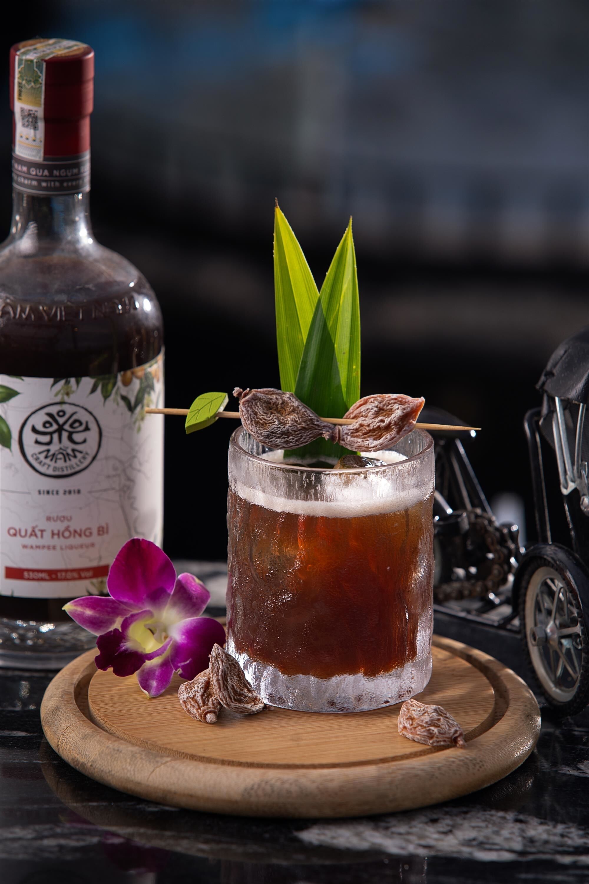 A glass of dark amber beverage with ice, garnished with a green leaf and a dried flower, on a wooden coaster. A bottle with a label is visible in the background.