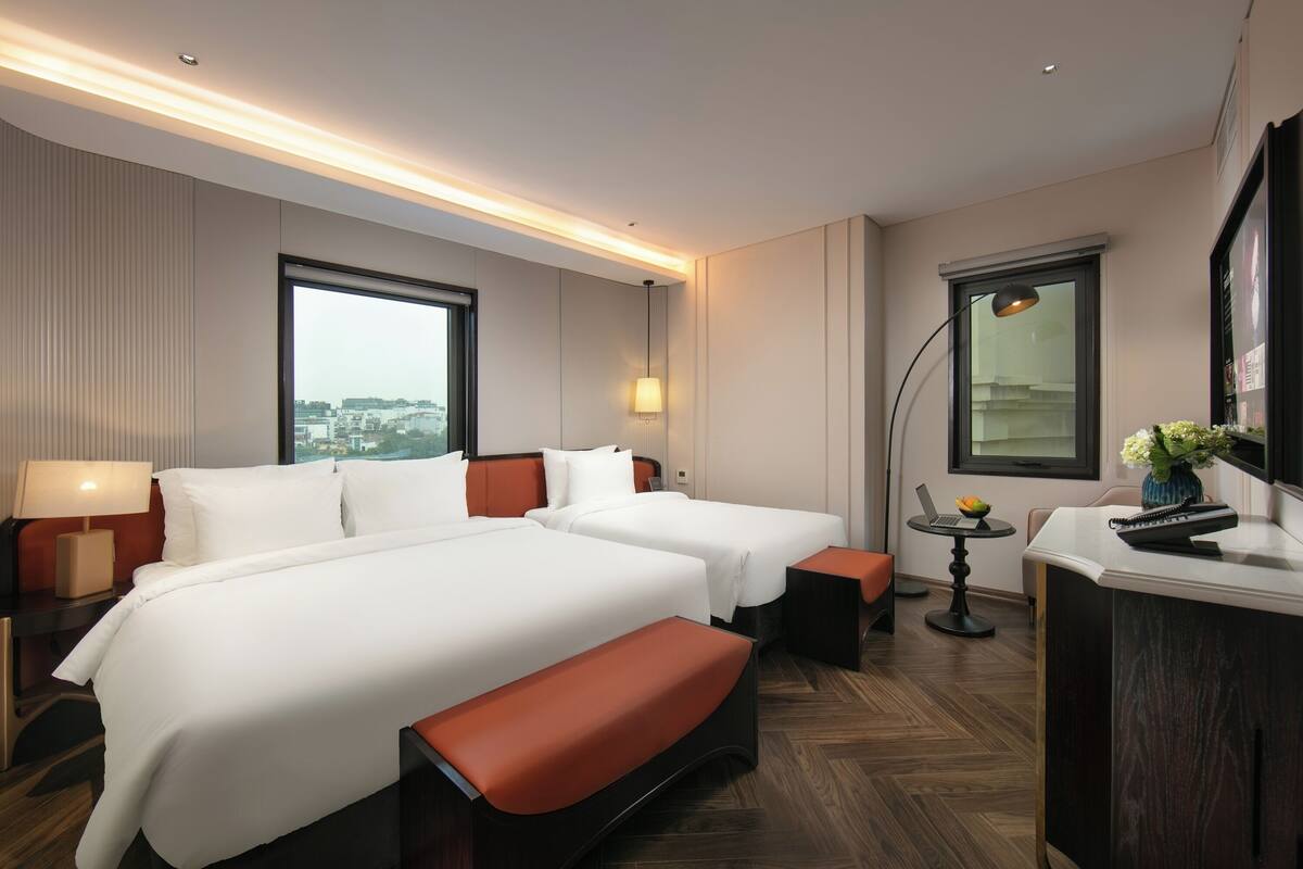 A hotel room with two beds, a desk, a chair, a lamp, and a window with a city view.
