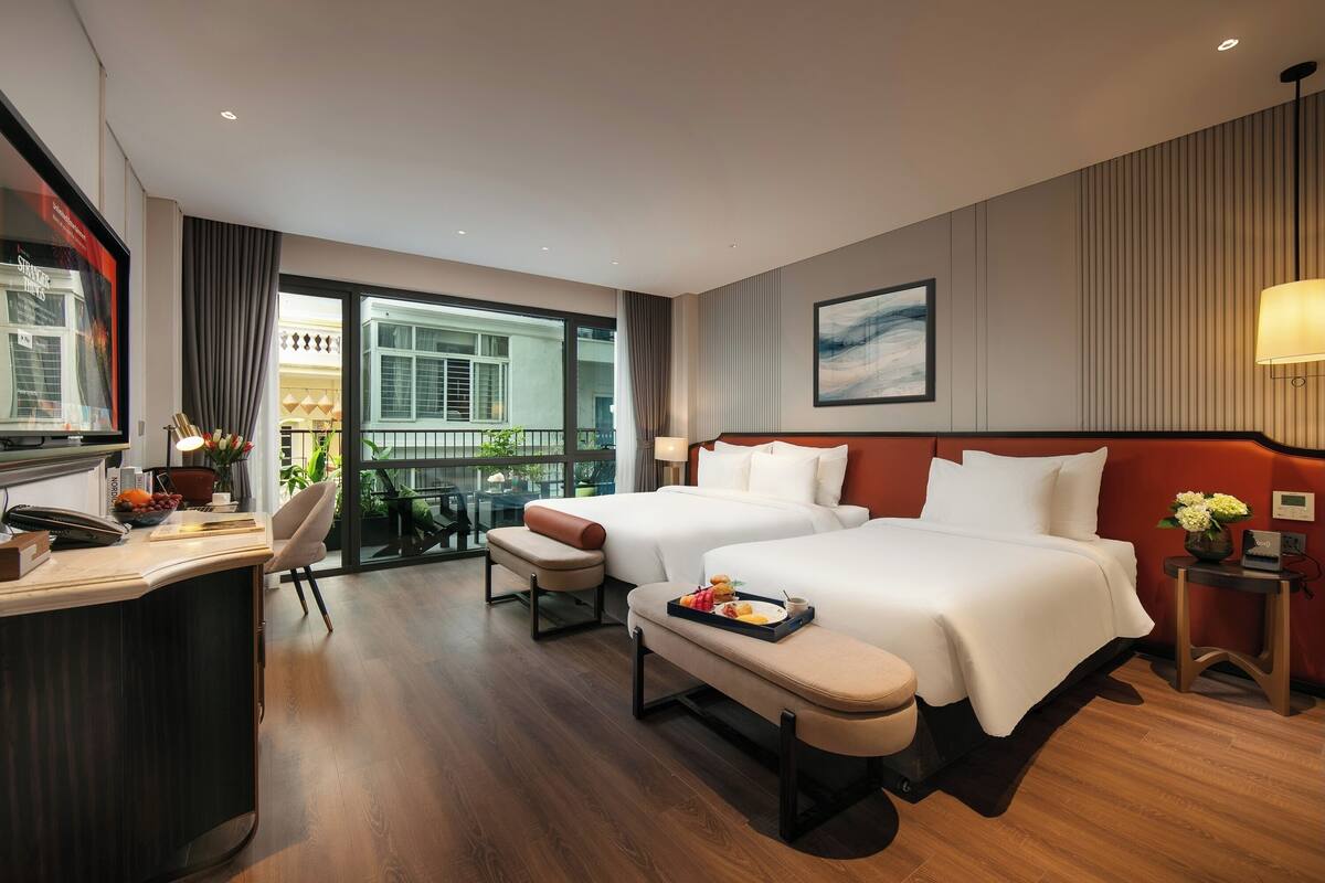 A hotel room with two beds, a desk with a telephone, a chair, a small table with flowers, and a view of the outside balcony.