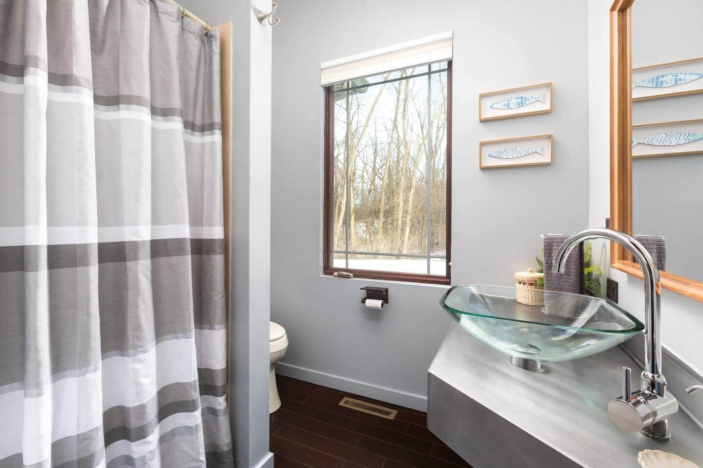 A bathroom with a glass sink, a toilet, a window with a view of trees, and a striped shower curtain.