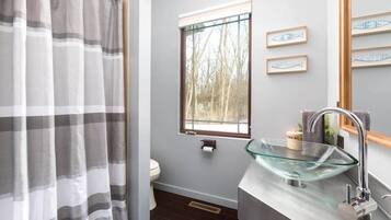 A bathroom with a glass sink, a toilet, a window with a view of trees, and a striped shower curtain.