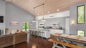 Un vaste espace de vie doté d’un plafond en bois, une cuisine avec des meubles blancs et un coin repas avec une table en bois.