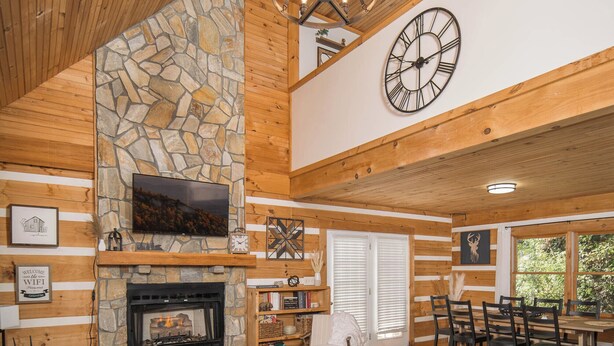 A cozy log cabin interior with a stone fireplace, wooden ceiling, and a dining area with a view of the outdoors. - Boone/Blowing Rock Cabin: Hot Tub, Mountain Views, Sleeps 8, Peaceful, Private (Vilas)