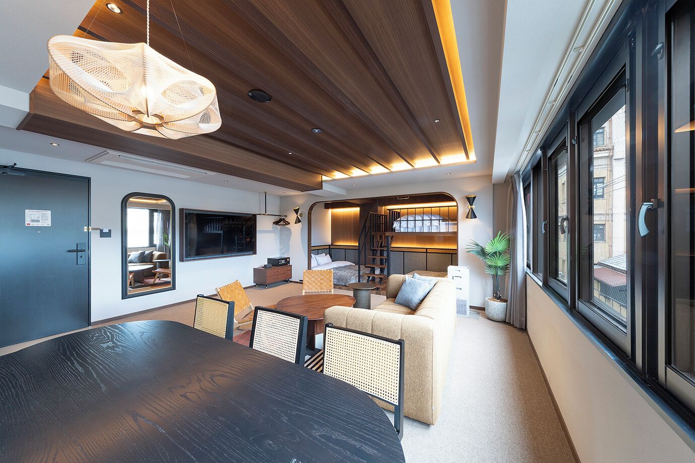 A modern hotel room with a dining area, a sofa, a large window, and a staircase leading to an upper level.