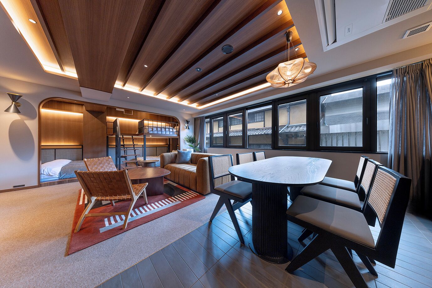 A modern hotel room with a wooden ceiling, a dining area with a round table and chairs, a sofa, and a view of the outside through large windows.