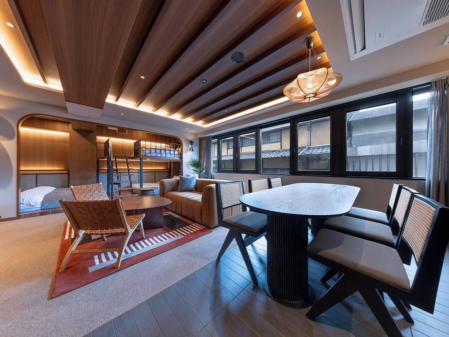 Una habitación de hotel moderna con techo de madera, una zona de comedor con una mesa redonda y sillas, un sofá y vistas al exterior a través de amplios ventanales.