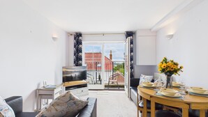 Ein modernes Wohnzimmer mit Fernseher, Sofa, Esstisch und Blick auf ein rotes Gebäude mit Ziegeldach durch eine Glastür.