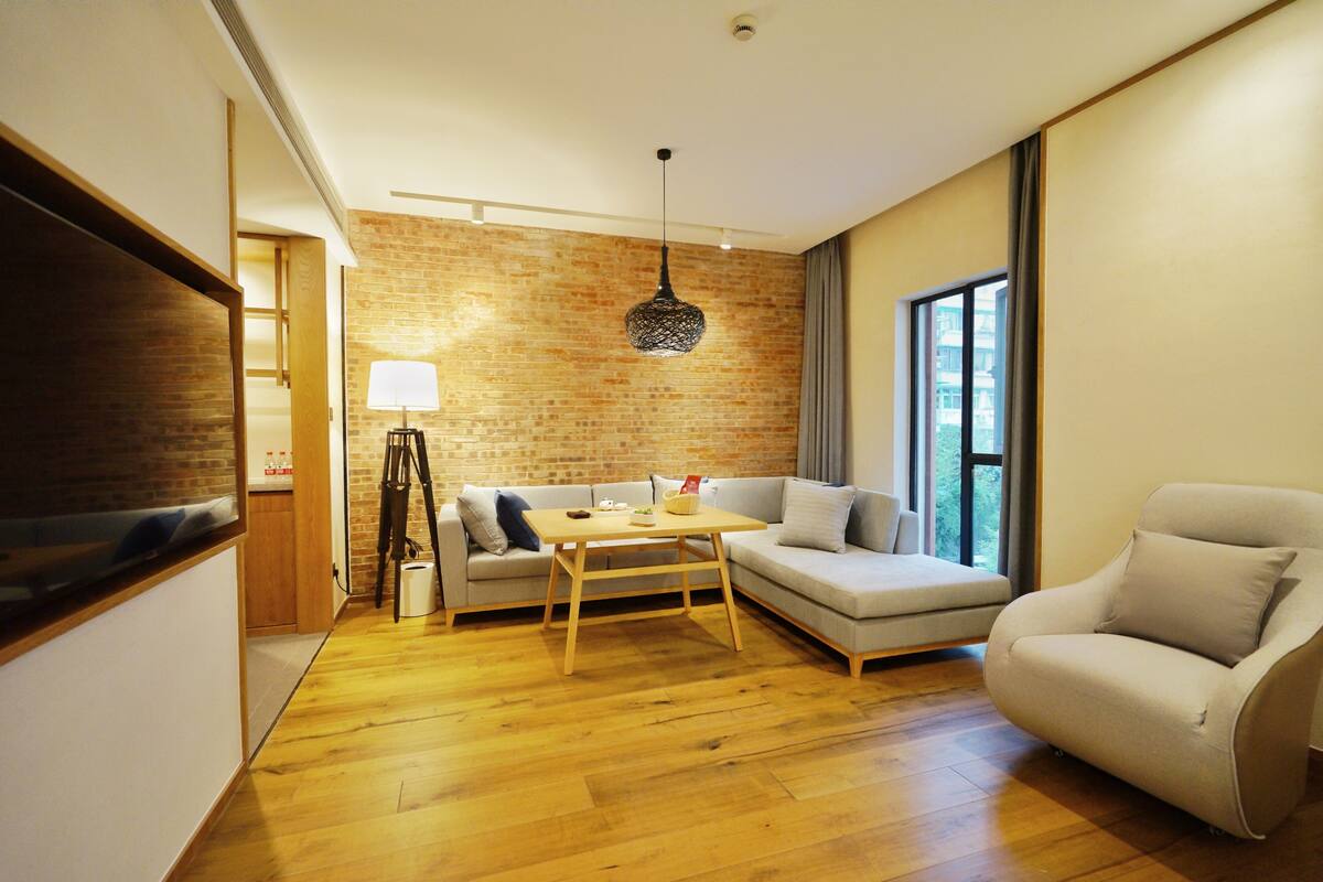 Un salon moderne avec un plancher en bois, un mur d’accent en briques, un canapé, un fauteuil, une table et un luminaire suspendu.