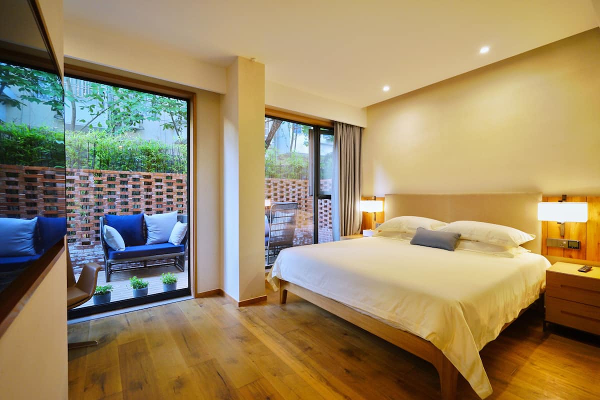 Une chambre avec un grand lit, un plancher en bois, un mur en briques et une vue sur un jardin, visible à travers une porte vitrée.