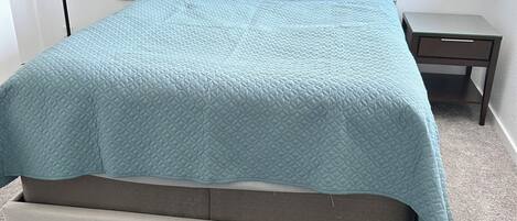 A bed with a quilted headboard, a small bedside table, and a large furry rug on the floor.