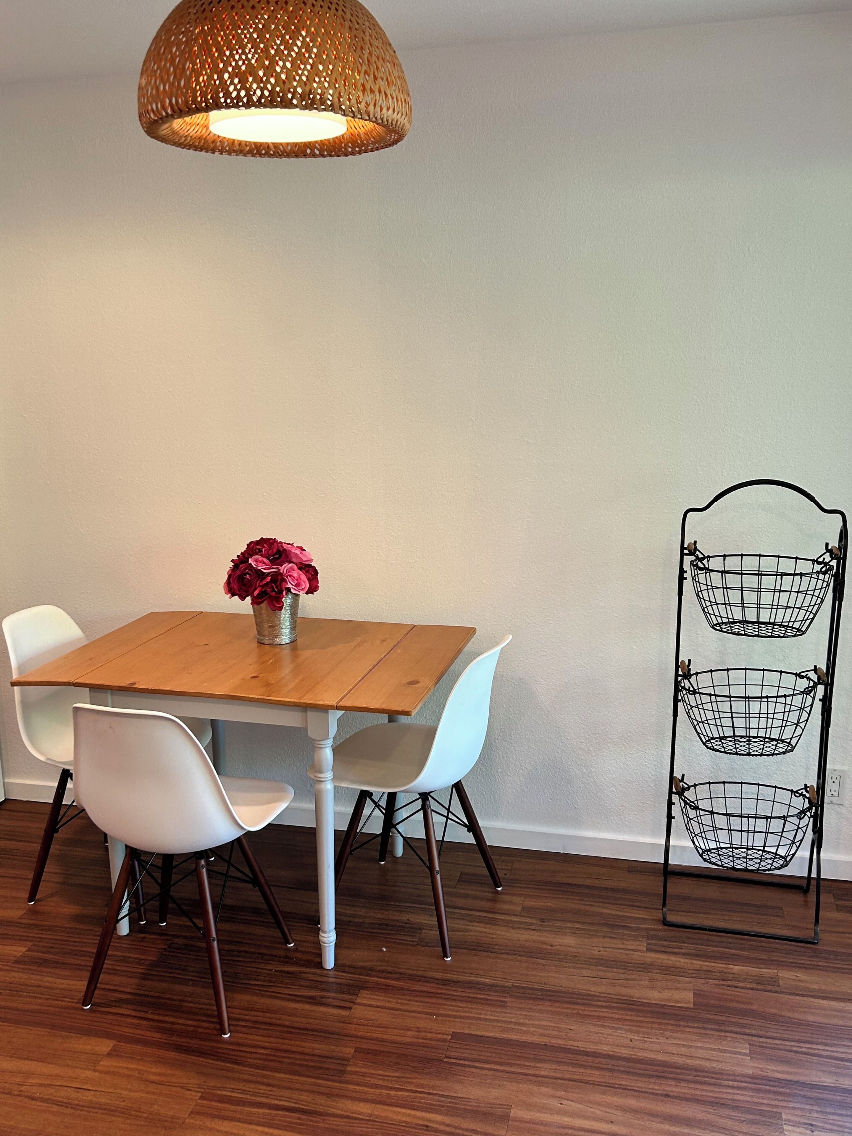 A dining area with a wooden table, white chairs, a hanging light fixture, and a metal rack against the wall.