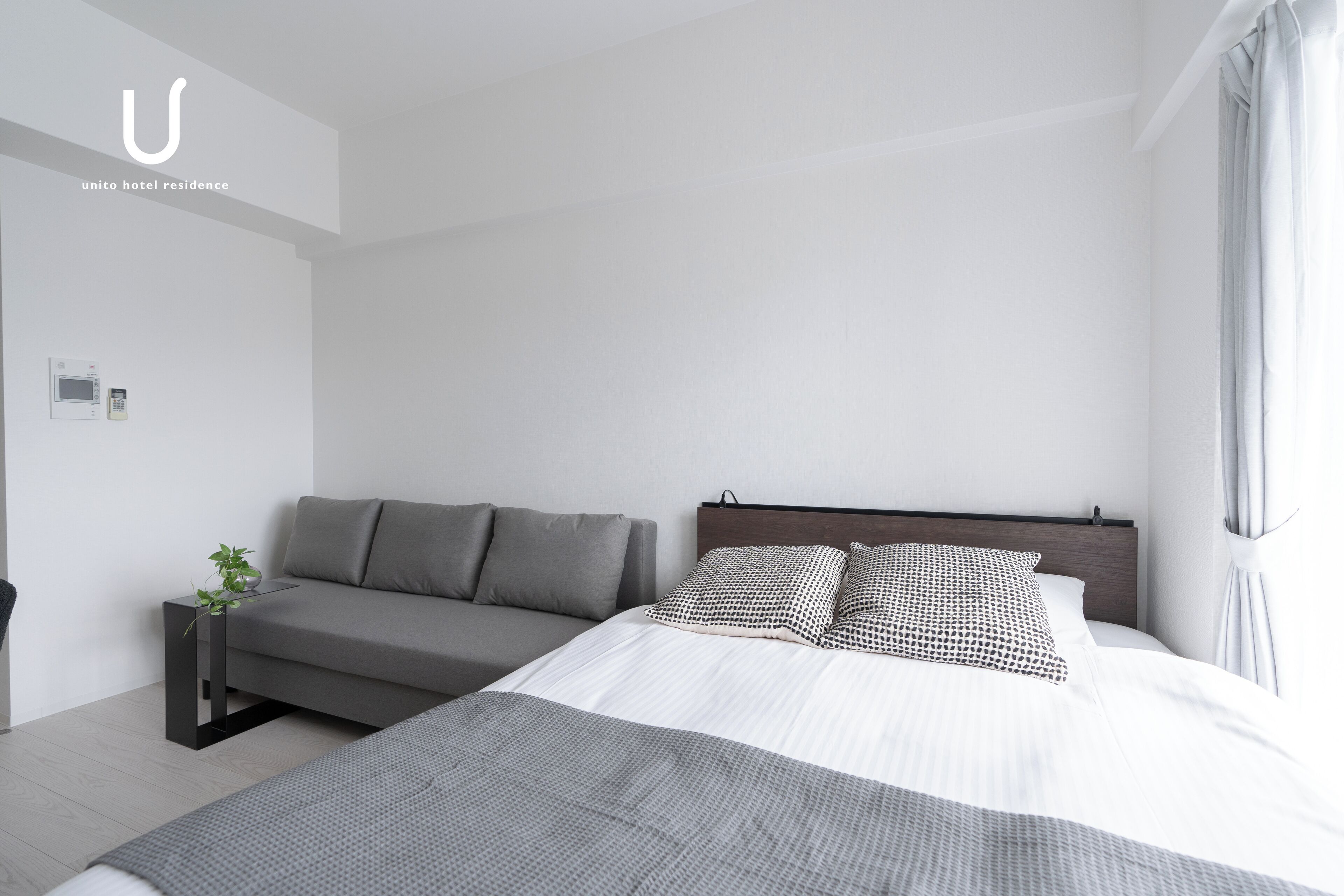 Una habitación de hotel moderna con una cama, un sofá, una mesita de noche con una planta y un panel de control en la pared.