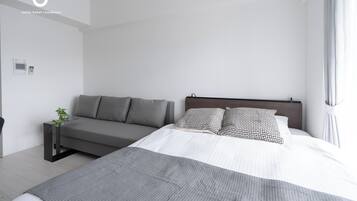 Una habitación de hotel moderna con una cama, un sofá, una mesita de noche con una planta y un panel de control en la pared.