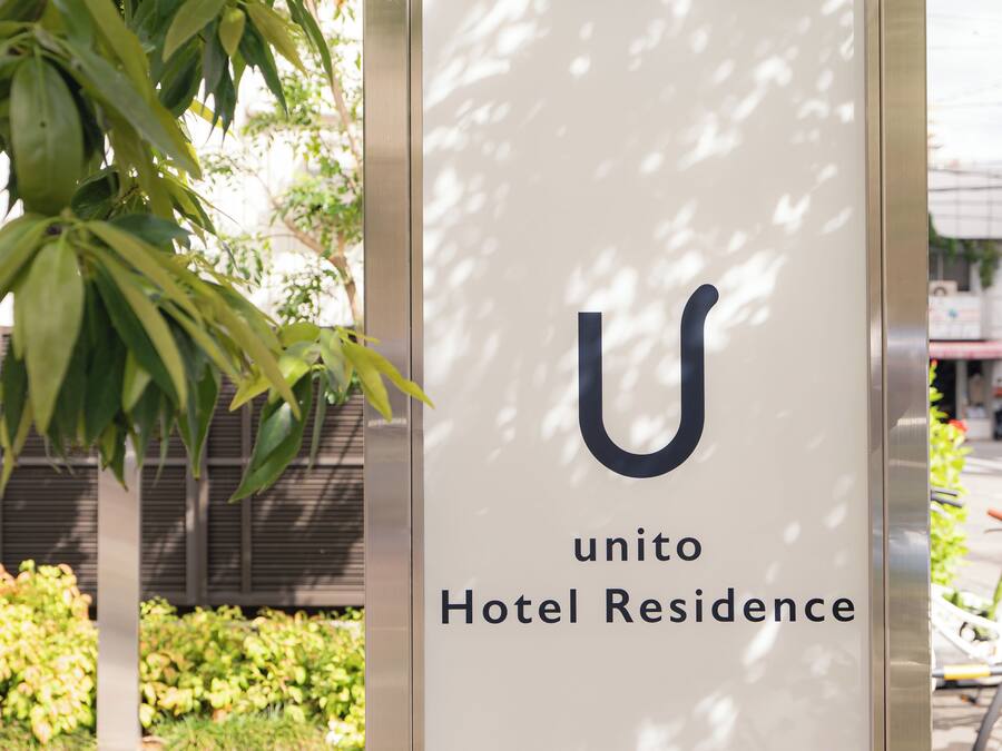 A sign for "unito Hotel Residence" with a large 'U' logo, surrounded by greenery and a building in the background.