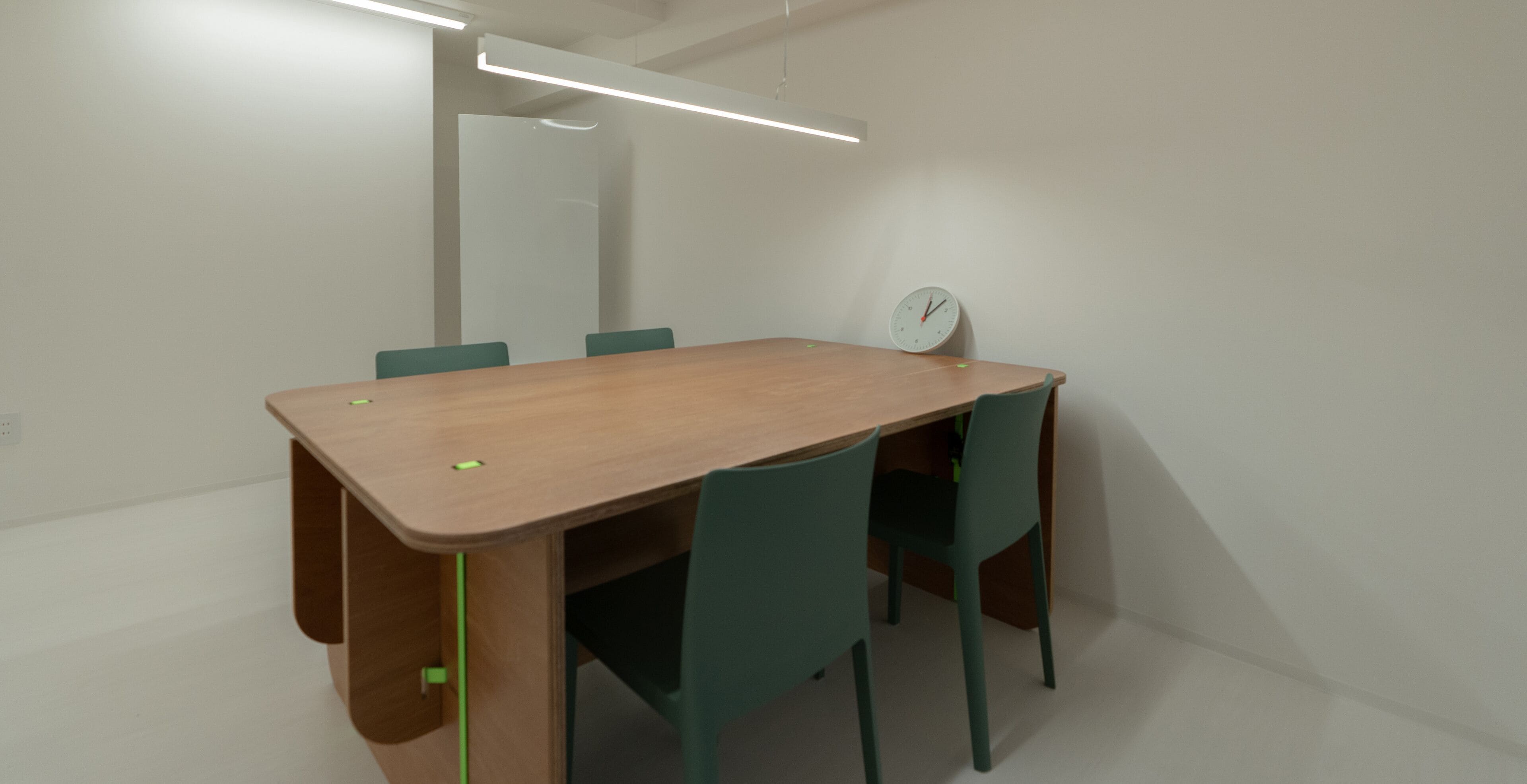 Une salle de conférence avec une table en bois, des chaises vertes, une horloge au mur et un luminaire moderne.
