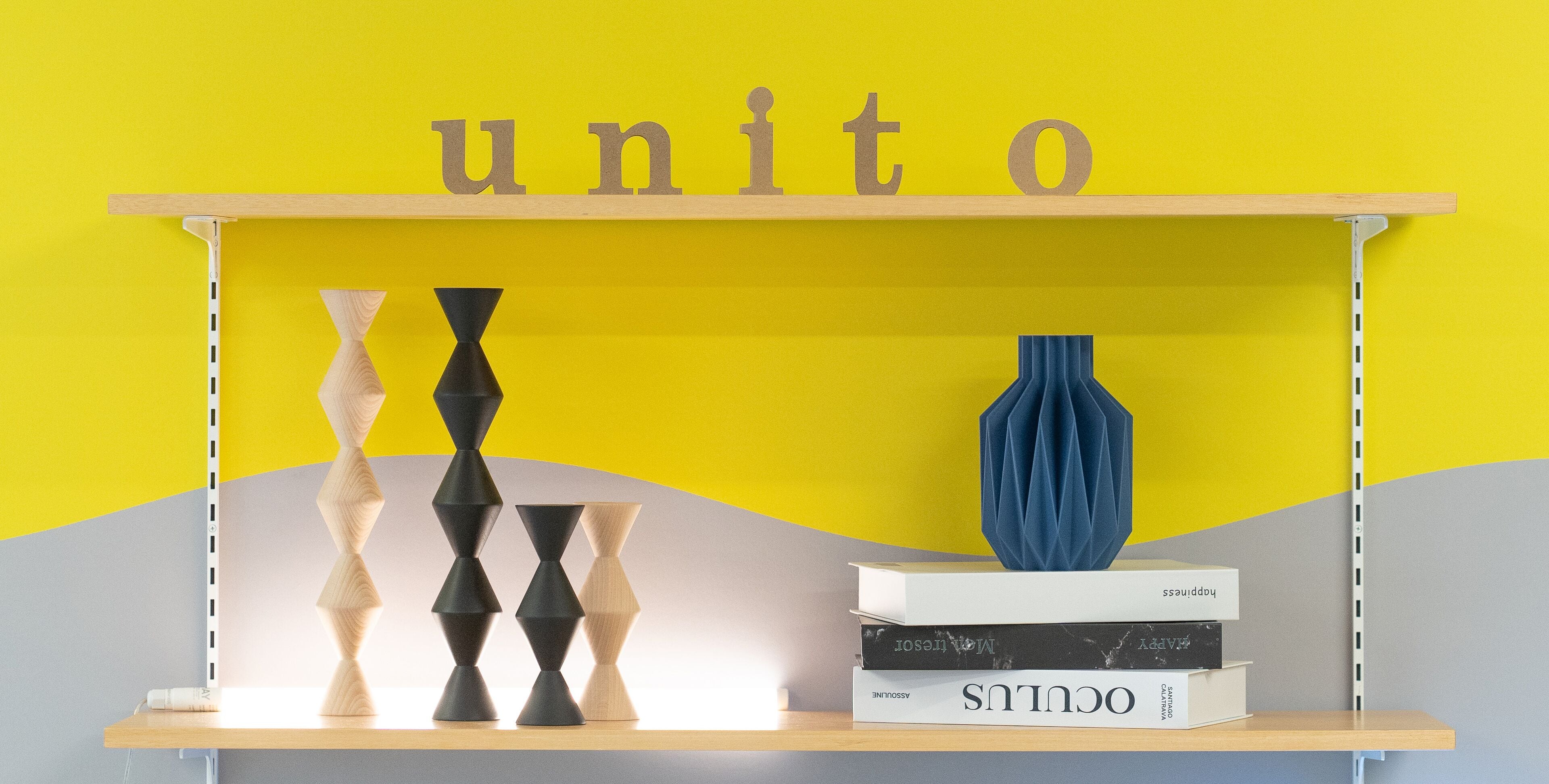 Una estantería con jarrones geométricos y libros frente a una pared amarilla con la palabra "unito" en la parte superior.