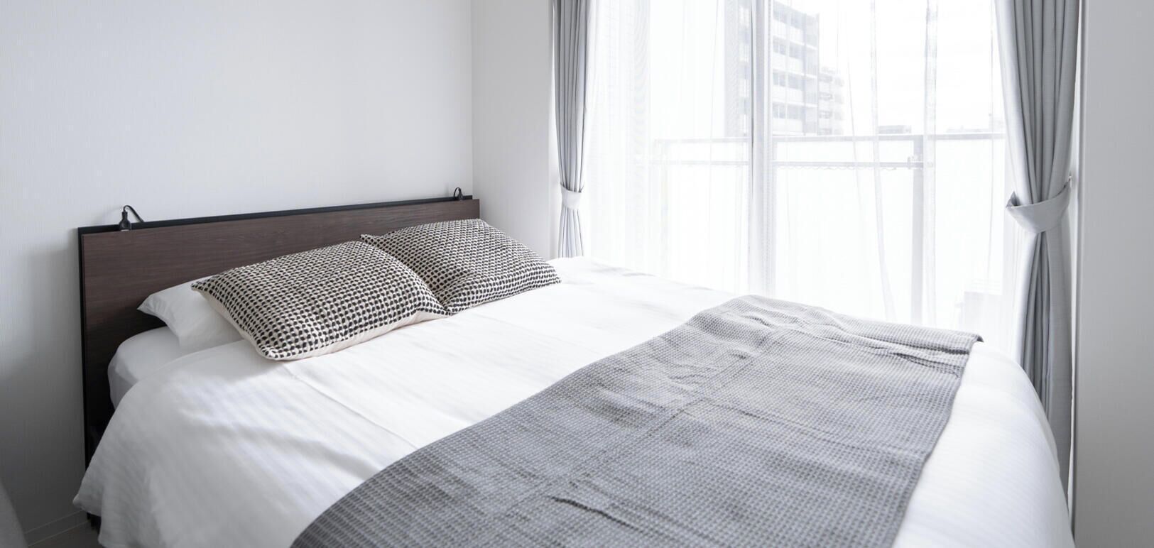 Een bed met een wit-grijs deken, twee gestreepte kussens en een donker hoofdbord in een kamer met een groot raam en gordijnen.