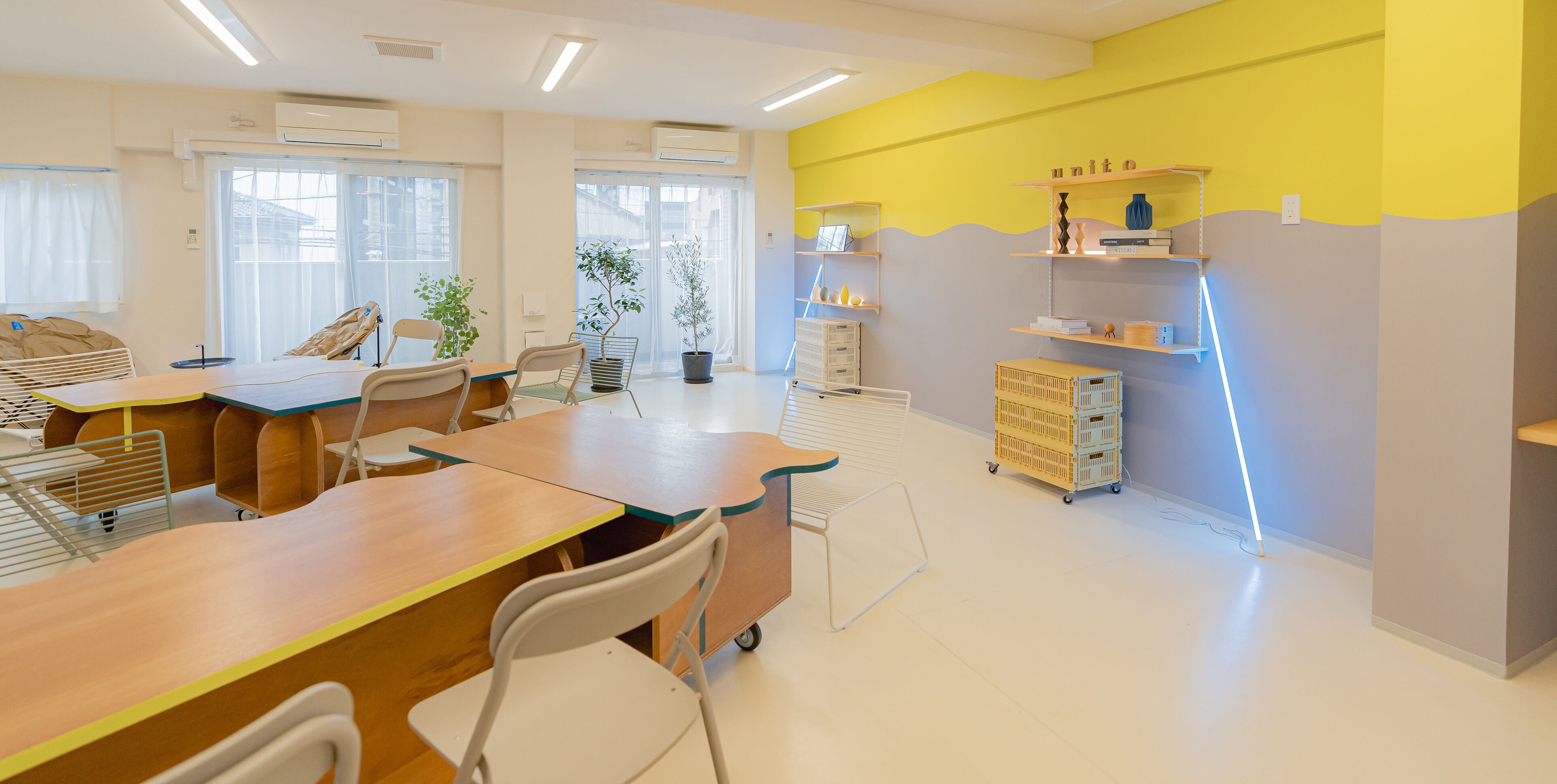 Una habitación moderna y luminosa con una amplia mesa de madera, varias sillas y un carrito. Hay plantas en macetas, estanterías con libros y objetos decorativos, y una pared de acento de color amarillo brillante.