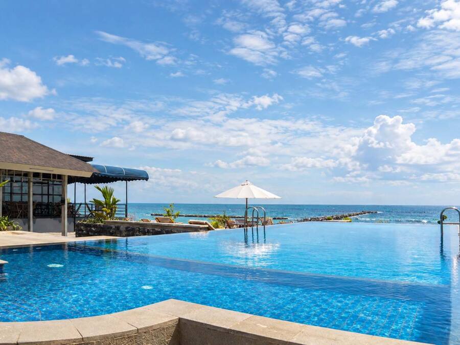 A clear blue pool with a view of the ocean, a covered seating area, and a lounge chair with an umbrella.