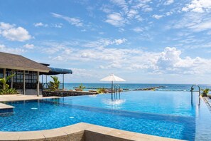 Une piscine d’un bleu éclatant avec vue sur l’océan, un espace salon abrité et un fauteuil de relaxation avec un parasol.