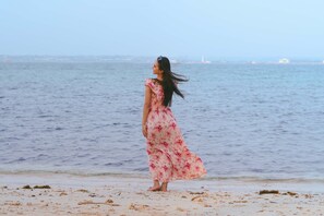 Une personne vêtue d’une robe à motifs floraux se promène sur une plage de sable, avec la mer et un phare lointain en arrière-plan.