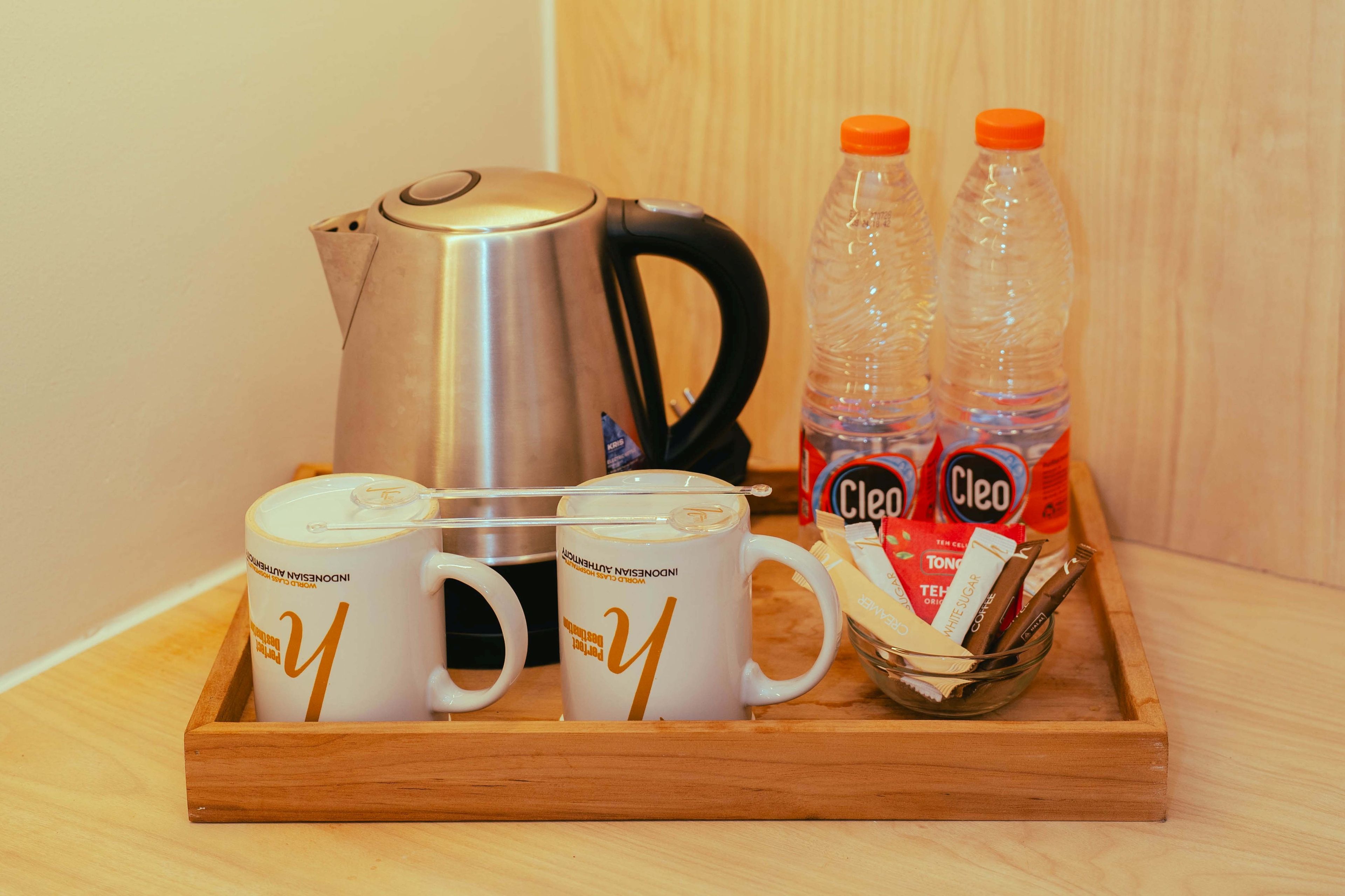 A tray with two white mugs, a stainless steel kettle, two plastic bottles, and a glass bowl containing a chocolate bar and a white chocolate bar.