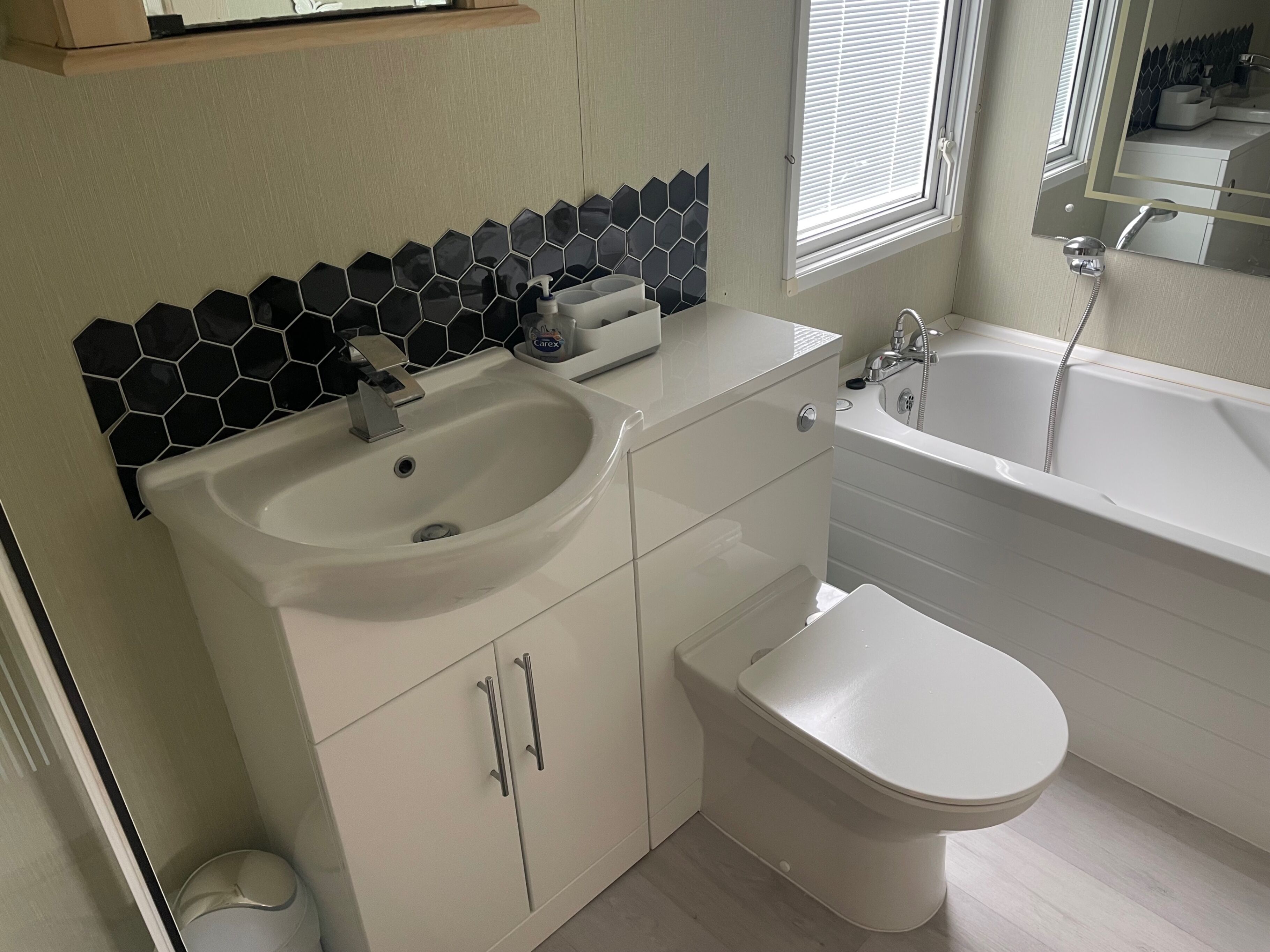 Un baño moderno con lavamanos, inodoro y bañera blancos. Cuenta con un respuesto de azulejos hexagonales negros sobre el lavamanos y una ventana con persianas en la pared.