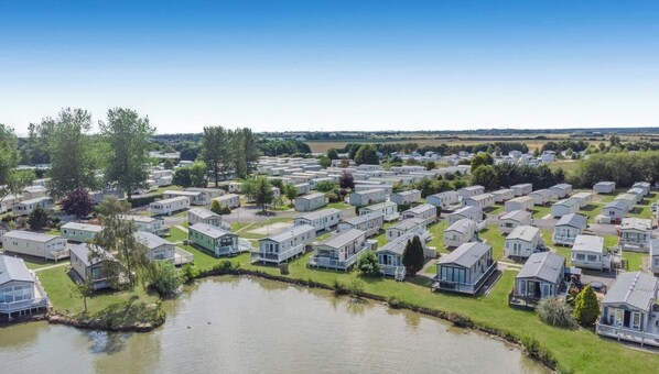 A large campground with numerous mobile homes arranged in rows, a small body of water in the foreground, and a clear blue sky.