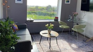 Ett modernt vardagsrum med en soffa, ett runt soffbord och en tv. Det finns ett fönster med utsikt över grönområden utanför.