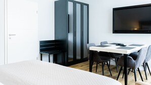 A modern hotel room with a bed, a dining table with chairs, a wardrobe, and a TV mounted on the wall.