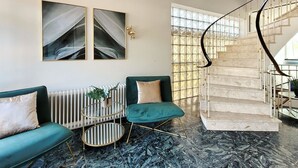 Una sala de estar moderna con un sofá verde, una mesa de centro redonda y un piso de mármol. Se observa una escalera con barandilla de vidrio al fondo.