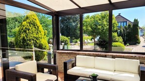 Un patio soleado con muebles de mimbre y una mesa de vidrio, con vistas a un jardín que tiene un árbol alto y una casa al fondo.