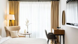 Habitación de hotel con cama, escritorio, silla, lámpara y un gran ventanal con cortinas.