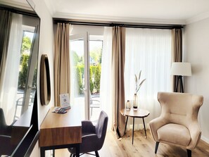 A hotel room with a wooden desk, a chair, a round table with a vase, a lamp, and a view of greenery through the window.