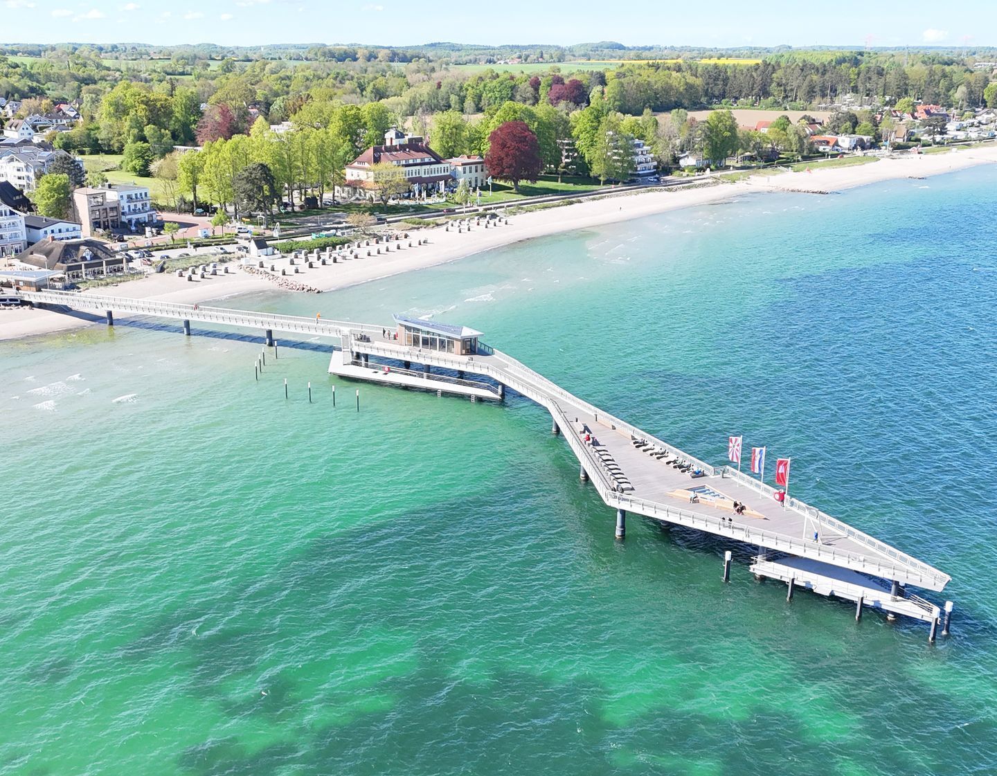 Une jetée s’étendant dans une étendue d’eau d’un bleu limpide, avec une plage et des bâtiments en arrière-plan.