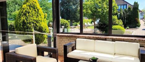 A sunlit patio with wicker furniture and a glass table, overlooking a garden with a tall tree and a house in the background.